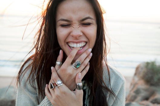 Girl smiling at the beach and biting her fingers with a lot of rings on at sunset.