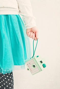 A girl in a turquoise tulle skirt, white cardigan with gold polka dots and black tights with white spots holds a retro mint camera at her side.