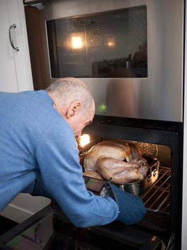 An older balding man reaches into the oven with large browning turkey.