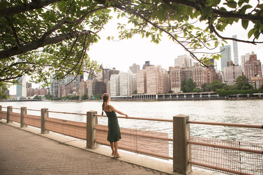 Young woman on Roosevelt Island leaning back from rail, looking out at the East river and NYC.