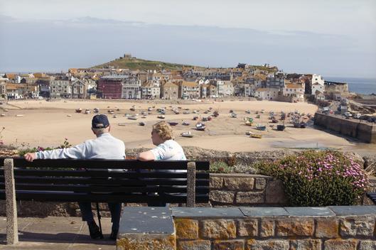 Two middle aged tourists sit on a bench lookking out over St. Ives harbour from a hill on a sunny day. In the distance are terraced houses, many of which are rented by holiday makers. The tide is out leaving the boats stranded on the sand. St. Ives, Cornw