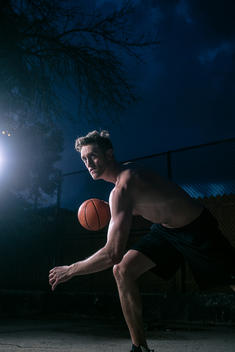 A male basketball player dribbling on a court at night while spotlit.