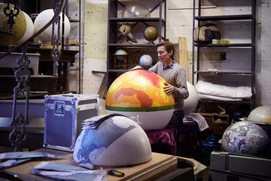 Man with hand holding large half decorated brightly lit globe in a dim workshop.