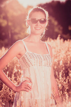 young woman with sunglasses standing at summer in a cornfield, wearing a light dress and smiling in camera, back light and lens flare