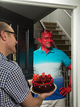 A man dressed as a devil stands with his front door open as his neighbor holds out a cake for him