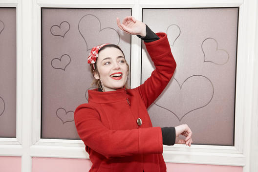 Young woman in a happy stance and red coat over a hearts background.