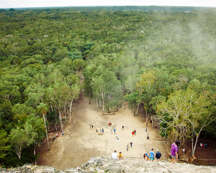 Visitors to a Mayan ruin climb its face whilst others congregate at its base. A large forest scales to the horizon