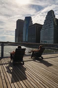 2 people in lounge chairs sitting in sun looking at manhattan
