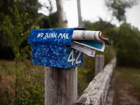 A blue outdoor mail box with No Junk Mail painted on it sits atop a fence post, completely full of junk mail.