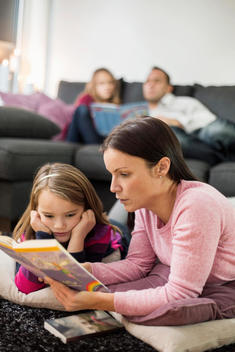 Mother and daughter reading story book on floor with family in background